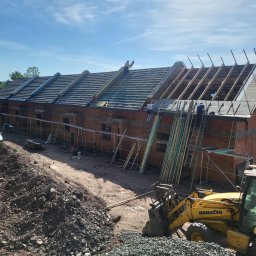 Grzegorz Kozicz - Budowa szeregowca z czerwonej cegły. Częściowo położony dach, widoczne rusztowania, drabiny i koparka Komatsu. Prace budowlane w toku, słoneczny dzień.