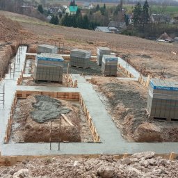 Grzegorz Kozicz - Zalane betonem fundamenty domu jednorodzinnego z widocznym zbrojeniem i szalunkiem. Na ziemi palety z bloczkami betonowymi. W tle pagórkowaty teren.