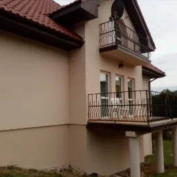 Nowo otynkowany dom jednorodzinny w Gorzuchowie z balkonami o metalowych, zdobionych balustradach i czerwoną dachówką, widok z boku.