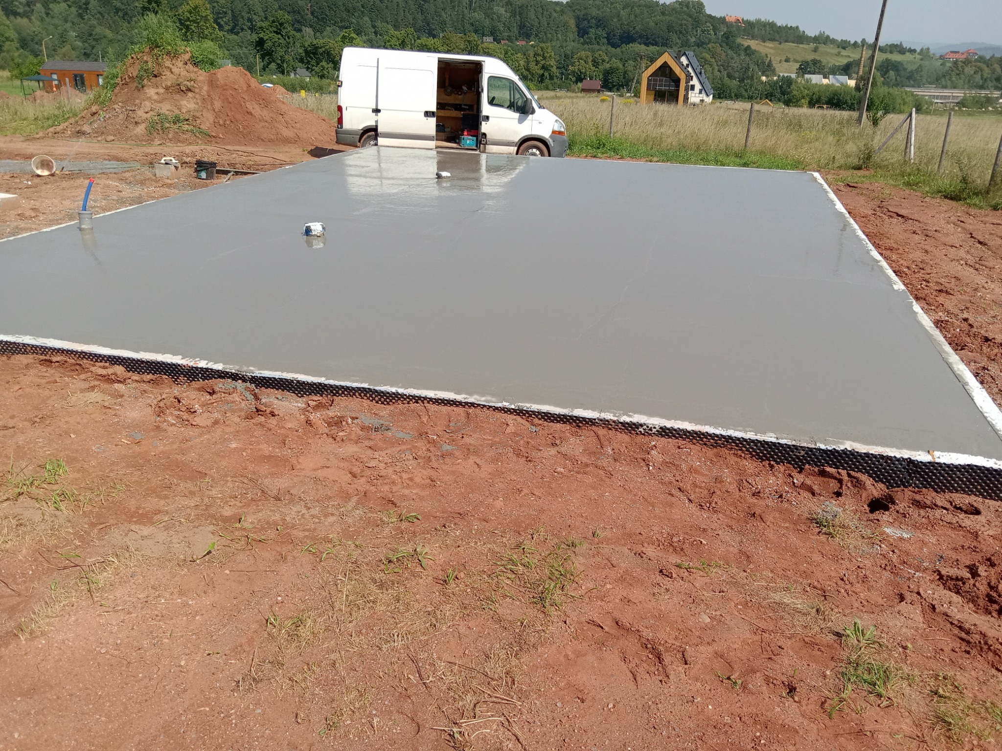 Świeżo wylana, gładka płyta fundamentowa z widoczną izolacją krawędziową, na tle samochodu dostawczego z otwartymi drzwiami i narzędziami w środku oraz zabudowy jednorodzinnej w oddali.