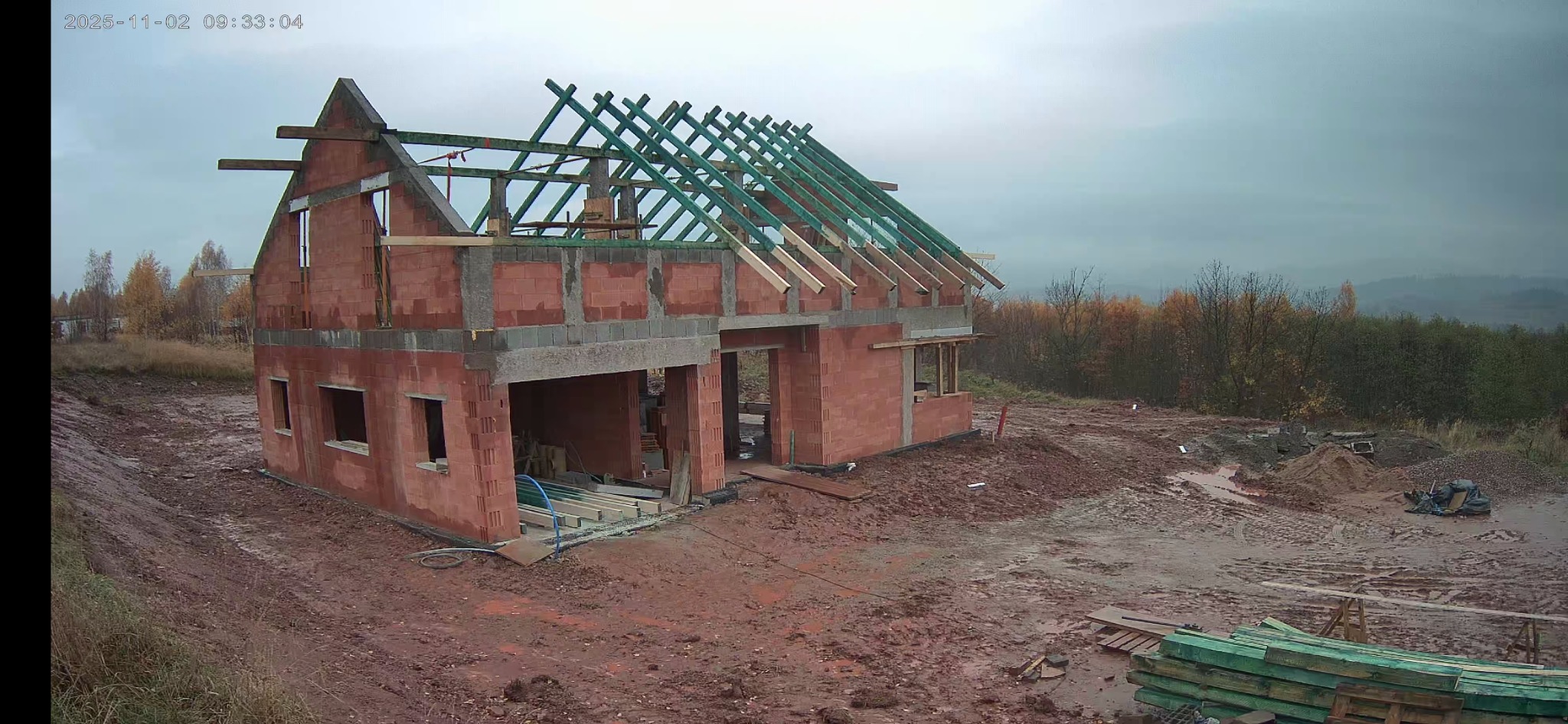 Dwukondygnacyjny dom w stanie surowym otwartym z czerwonej cegły, z widoczną konstrukcją dachu i elementami szalunków, na tle jesiennego krajobrazu i pochmurnego nieba.