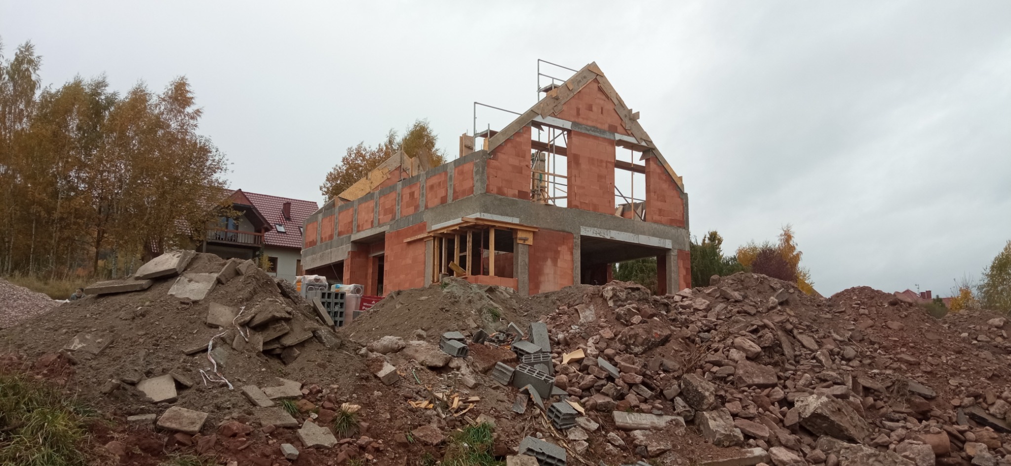 Surowy stan domu z czerwonej cegły w trakcie budowy, z widocznymi elementami konstrukcyjnymi i stertą gruzu na pierwszym planie, na tle pochmurnego nieba.