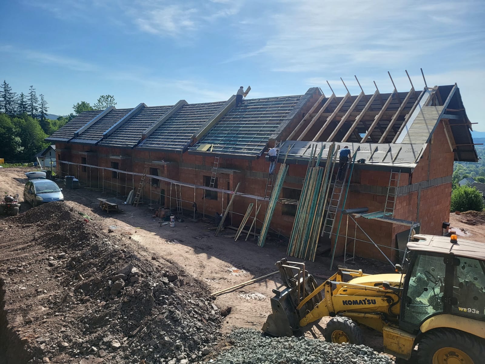 Budowa szeregowca z czerwonej cegły. Częściowo położony dach, widoczne rusztowania, drabiny i koparka Komatsu. Prace budowlane w toku, słoneczny dzień.