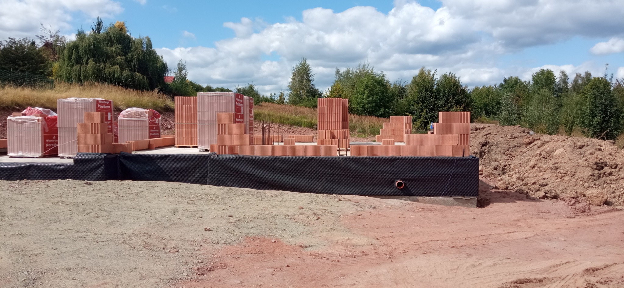 Budowa fundamentów z bloczków Porotherm w Gorzuchowie. Widoczne zabezpieczenie przeciwwilgociowe i przygotowane materiały budowlane. Ujęcie w słoneczny dzień.