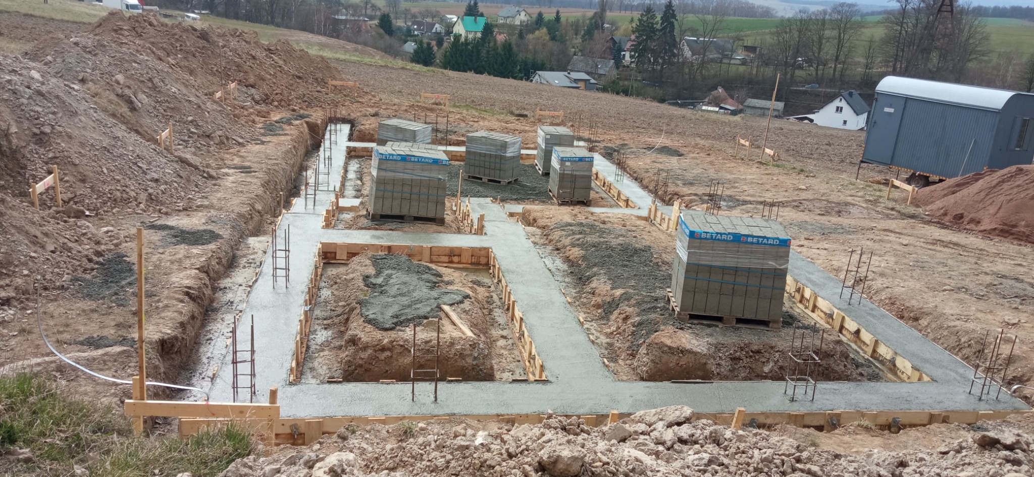 Zalane betonem fundamenty domu jednorodzinnego z widocznym zbrojeniem i szalunkiem. Na ziemi palety z bloczkami betonowymi. W tle pagórkowaty teren.
