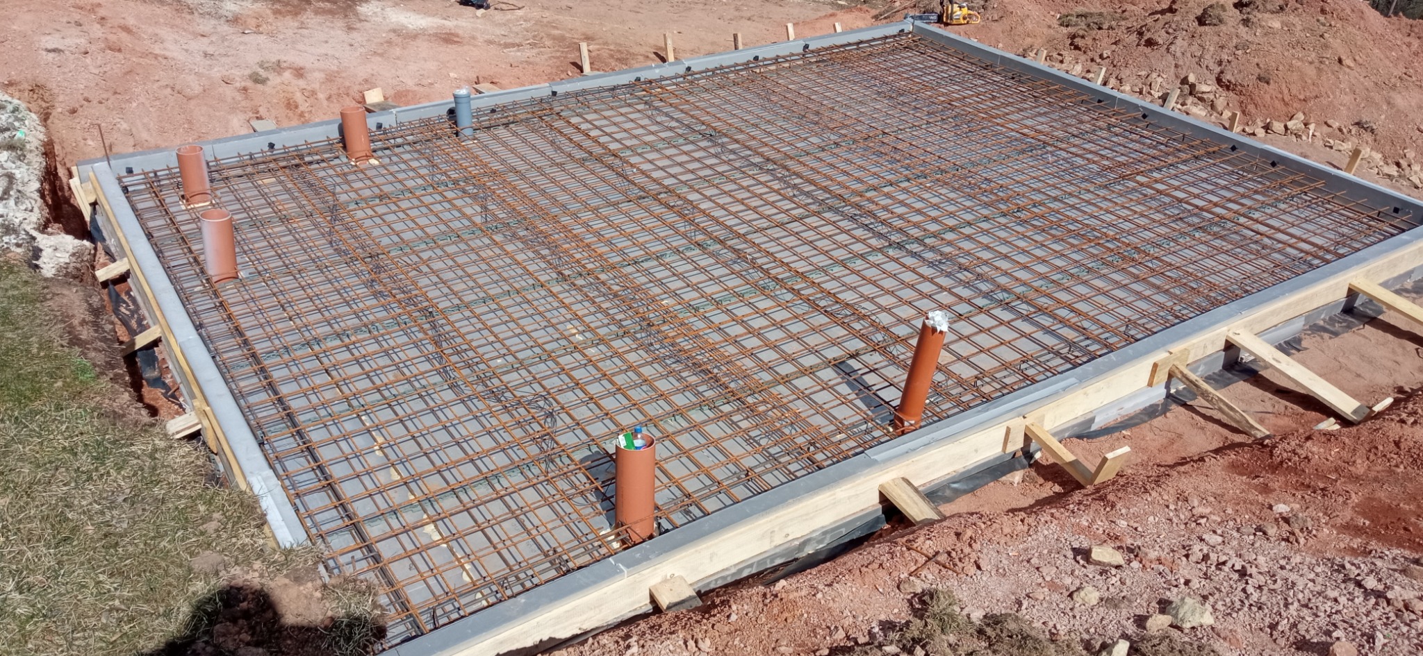 Zbrojenie płyty fundamentowej z rurami kanalizacyjnymi, widok z góry. Stalowa siatka na betonowej wylewce w szalunku, przygotowana do zalania betonem.