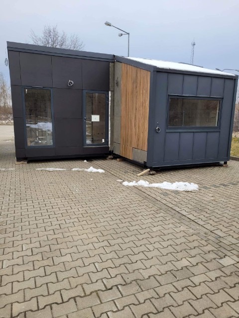 Dwa moduły nowoczesnego budynku z szarej blachy i elementami drewnopodobnymi, ustawione na kostce brukowej, z widocznymi fragmentami śniegu na dachu i kostce.