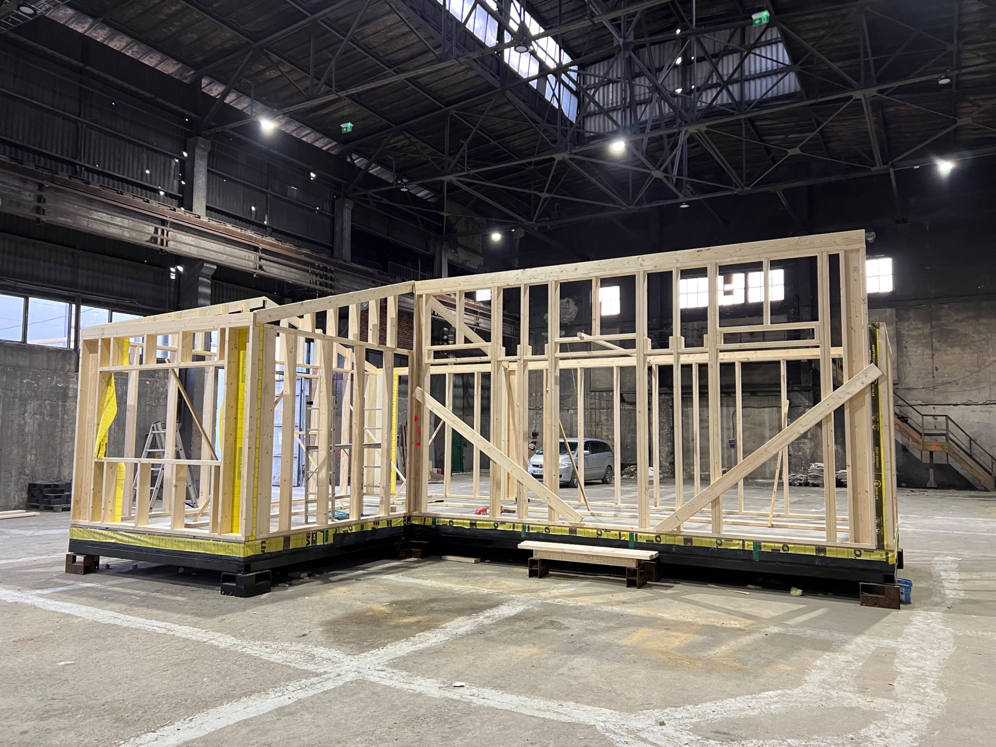 Wnętrze hali z częściowo zmontowaną konstrukcją szkieletową domu na czarnej platformie, widoczny samochód w tle i metalowe schody.