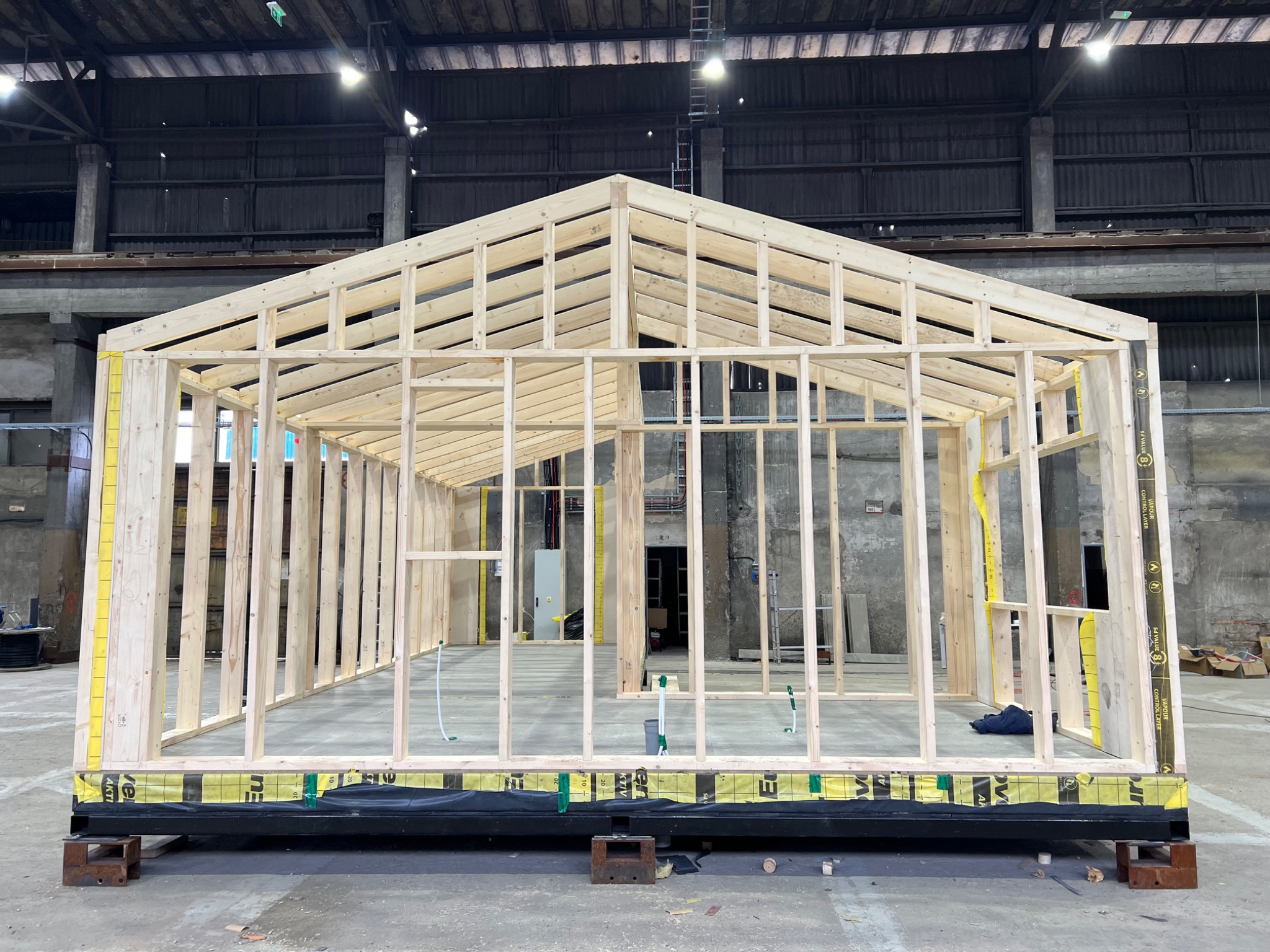 Konstrukcja szkieletowa budynku w hali produkcyjnej, widoczne elementy drewniane i izolacja, surowy beton w tle.