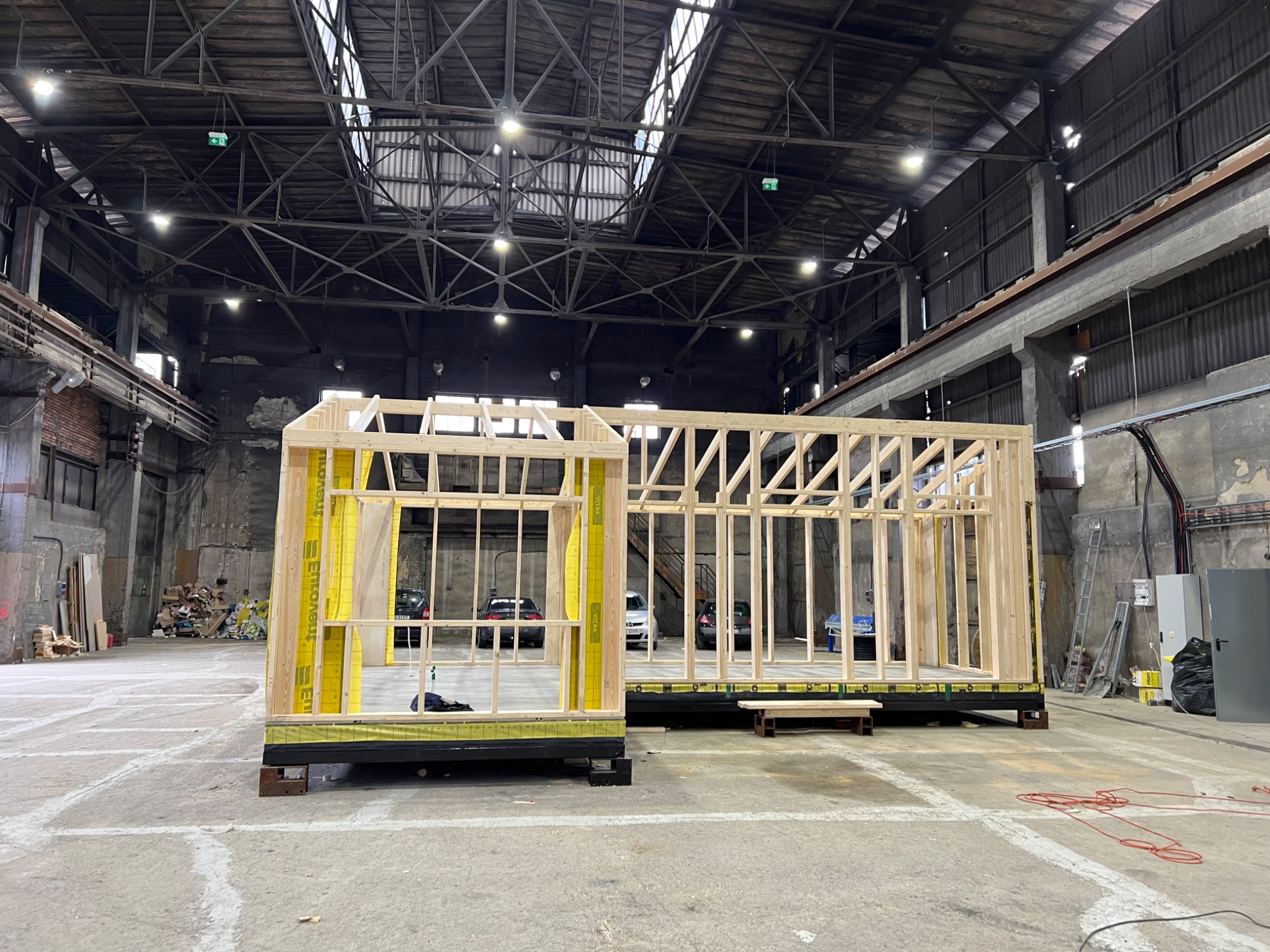 Dwa moduły domu szkieletowego w trakcie budowy, z widoczną konstrukcją drewnianą i żółtą izolacją, umieszczone w przestronnej hali przemysłowej z widocznymi elementami konstrukcyjnymi dachu.