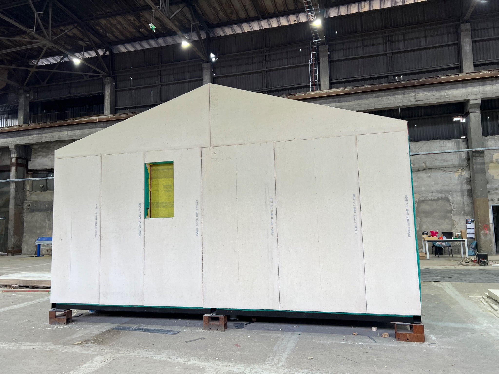 Wnętrze hali produkcyjnej z widocznym fragmentem prefabrykowanej ściany domu z otworem okiennym, opartej na drewnianych klockach.