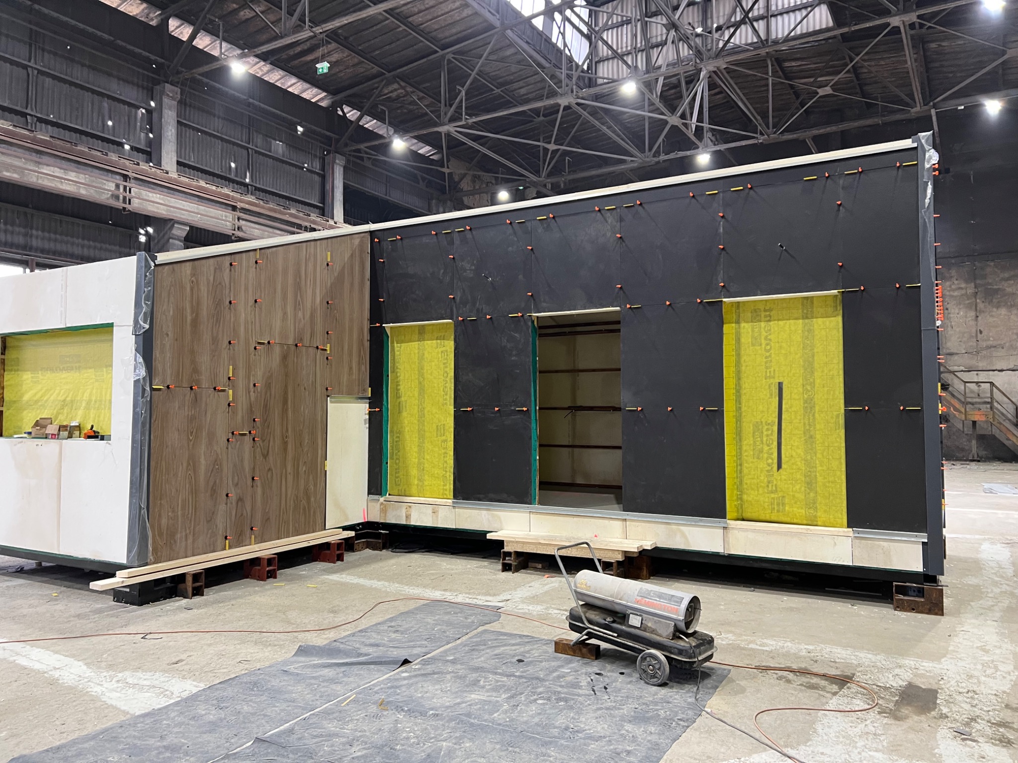 Moduł domu w trakcie budowy w hali produkcyjnej, widoczne warstwy izolacji, drewniana okładzina i nagrzewnica powietrza.