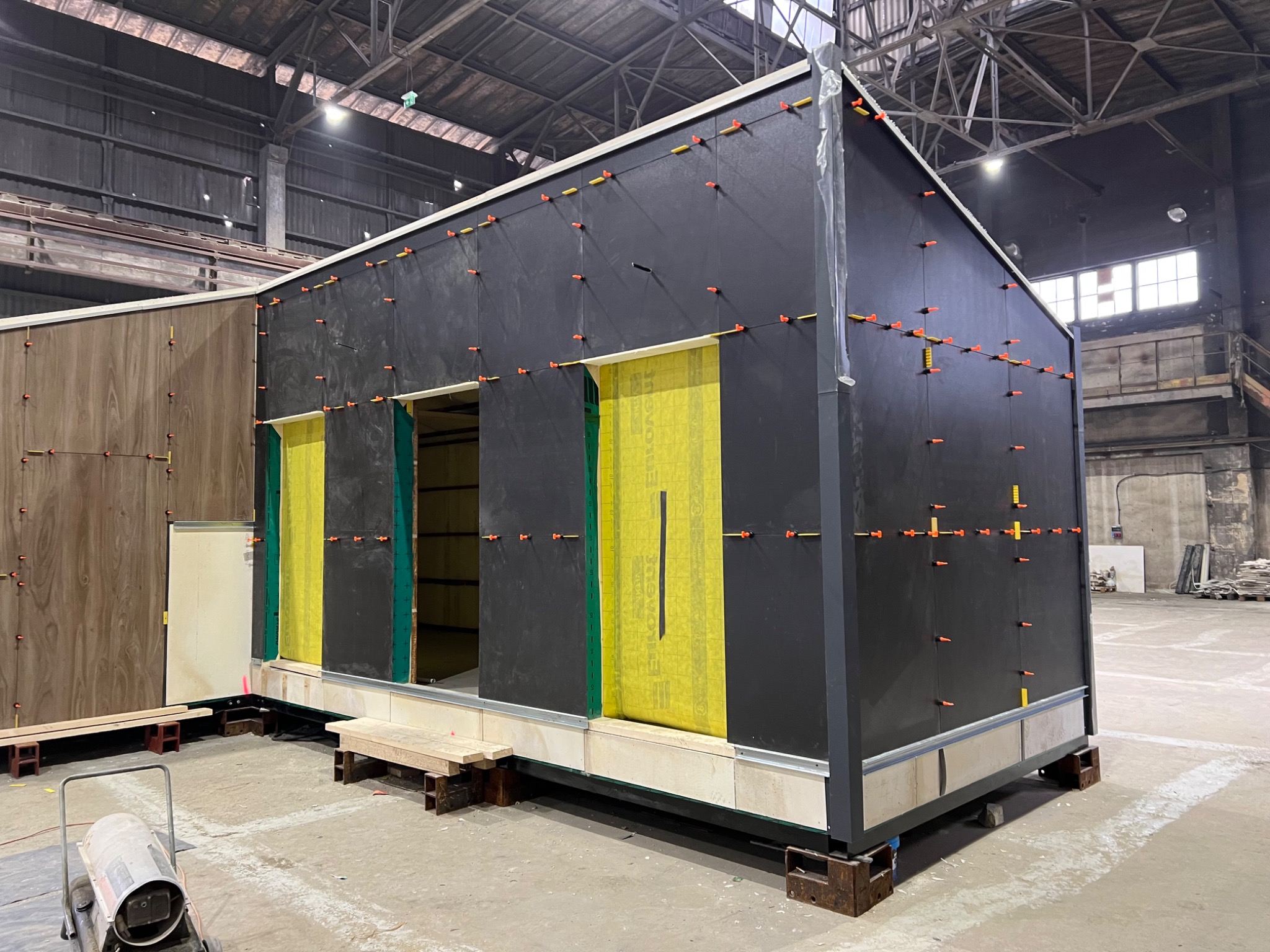 Fragment prefabrykowanego modułu z czarną elewacją, żółtą izolacją w otworach okiennych i drzwiowych, mocowany za pomocą pomarańczowych kołków, widoczny w hali produkcyjnej.