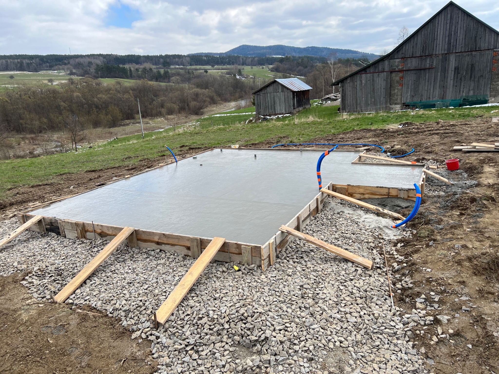 Wylany fundament pod budynek z drewnianym szalunkiem, widoczne niebieskie rury instalacyjne, w tle zabudowania gospodarcze i pagórkowaty krajobraz.