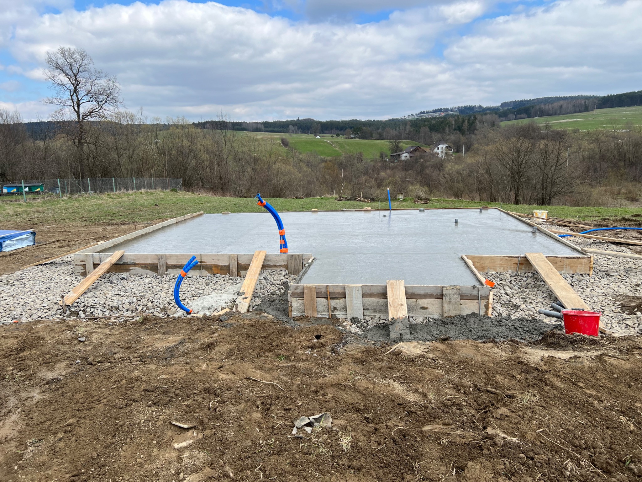 Wylana płyta fundamentowa z drewnianym szalunkiem i wystającymi rurami instalacyjnymi na tle pagórkowatego krajobrazu.