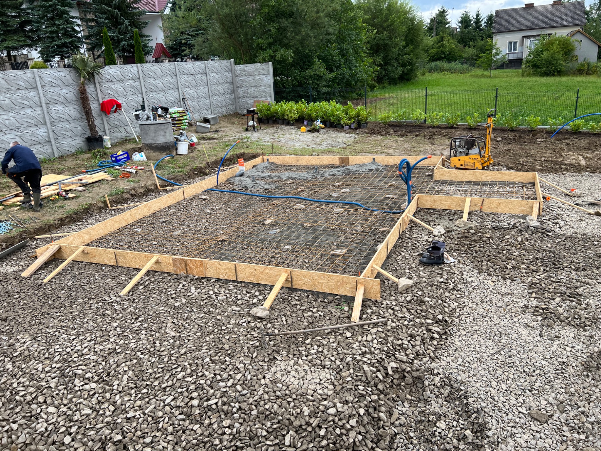 Przygotowanie fundamentów pod dom szkieletowy: szalunek z drewnianych desek, zbrojenie stalowe, ubijarka mechaniczna, widoczna instalacja wodna, teren wysypany żwirem, w tle dom jednorodzinny...