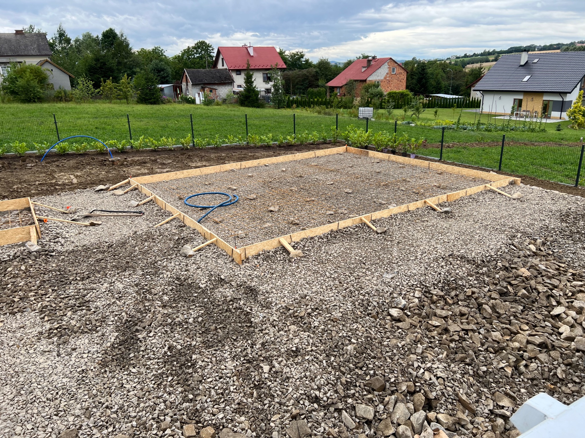 Przygotowanie podłoża pod budowę: szalunek z desek, siatka zbrojeniowa na żwirze, widok na domy w tle.