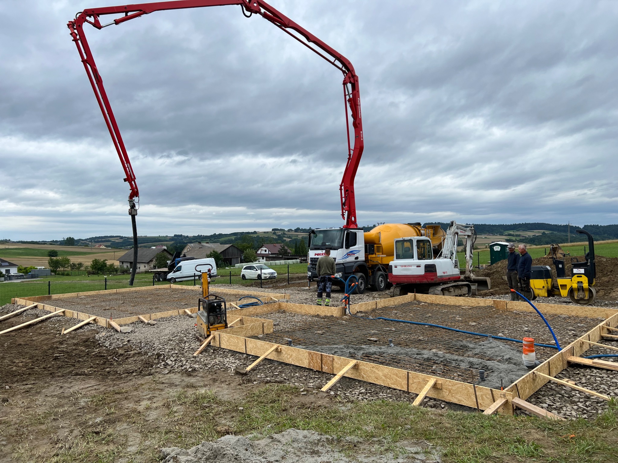 Wylewanie betonu na zbrojenie fundamentów pod budowę, widoczna pompa do betonu, mini koparka i zagęszczarka, w tle zabudowa wiejska.