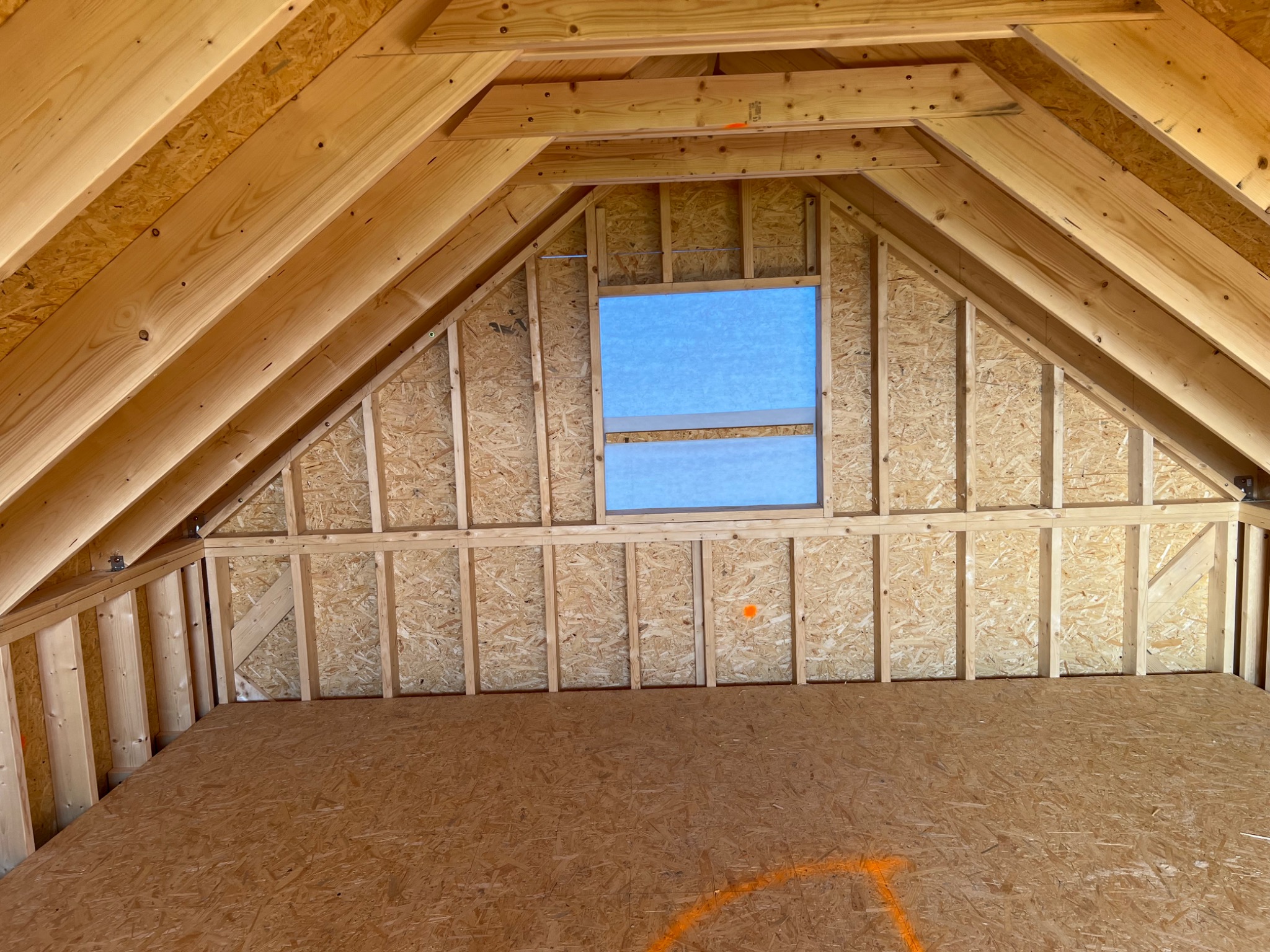 Wnętrze domu szkieletowego w trakcie budowy, widoczne drewniane elementy konstrukcyjne ścian, dachu i podłogi z płyt OSB, okno zabezpieczone niebieską folią.