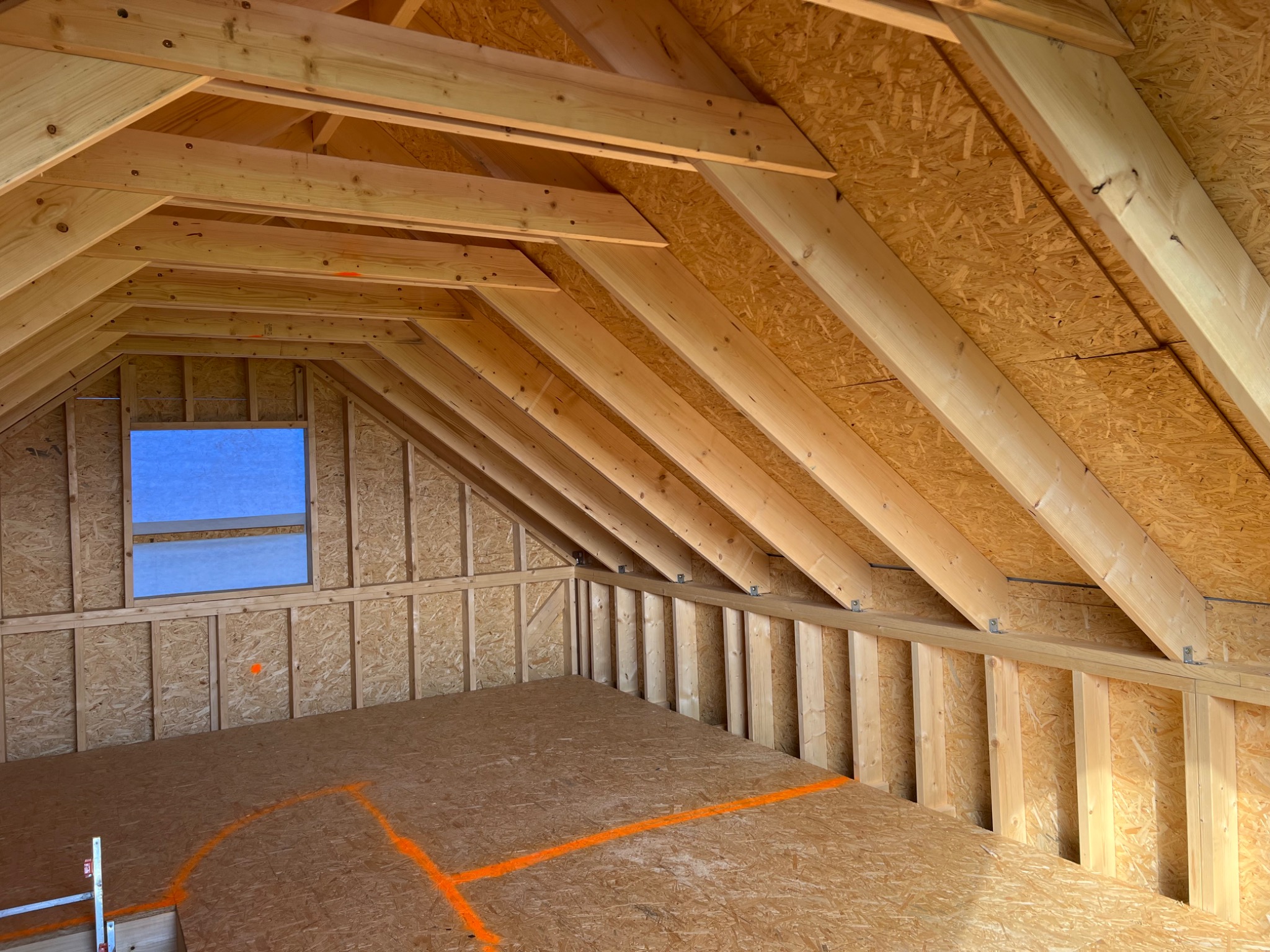 Wnętrze w trakcie budowy: szkielet dachu z widocznymi belkami, okno z niebieską folią ochronną, podłoga z płyt OSB z pomarańczowymi oznaczeniami