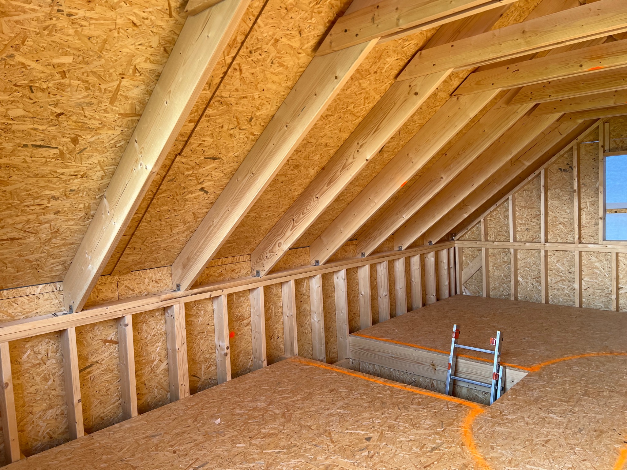 Wnętrze poddasza w trakcie budowy, widoczne drewniane belki konstrukcyjne, płyty OSB, małe okno i aluminiowa drabinka.