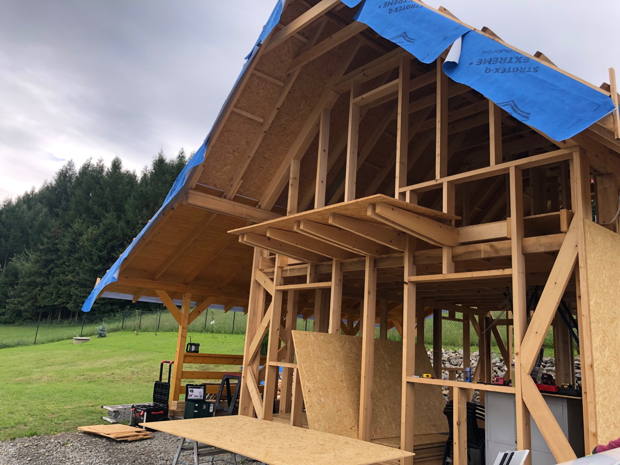 Konstrukcja szkieletowa domu w trakcie budowy, widoczne drewniane elementy i niebieska membrana dachowa Strotex-Q Extreme, narzędzia na placu budowy.