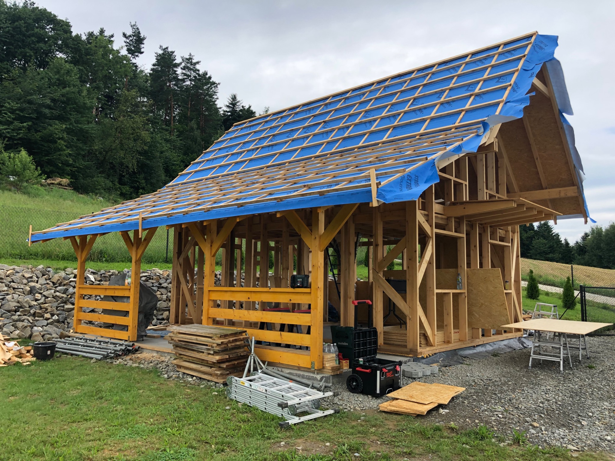 Konstrukcja w trakcie budowy z drewnianych belek, zabezpieczona niebieską membraną dachową, w tle zalesione wzgórze.