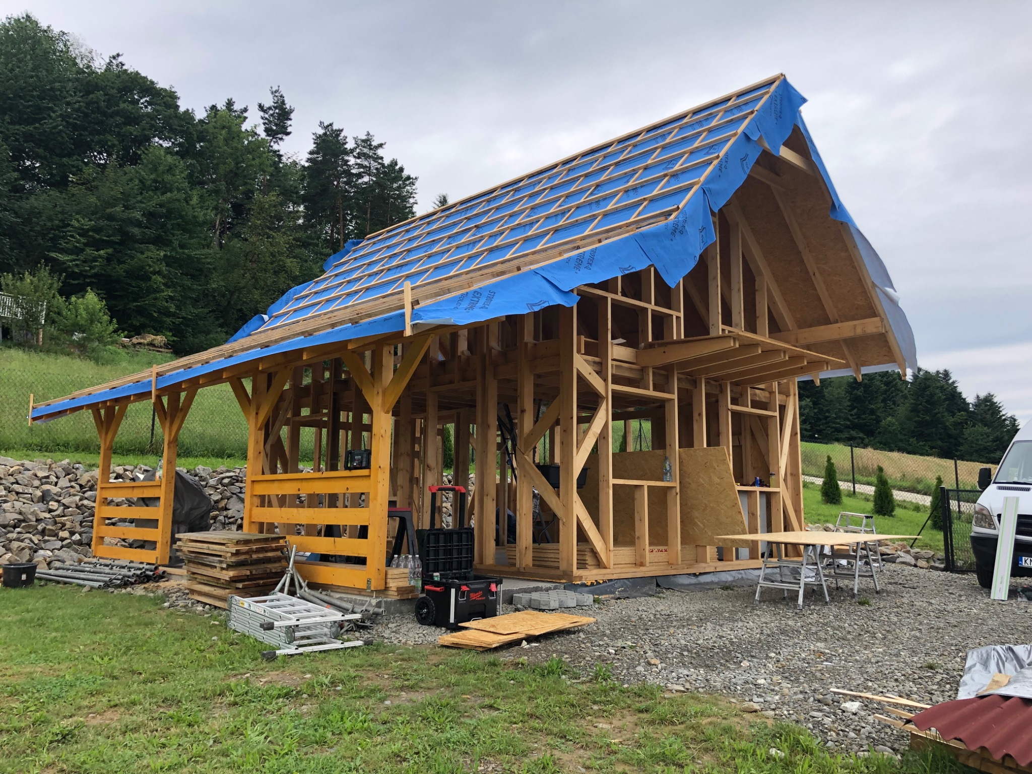 Konstrukcja szkieletowa domu w trakcie budowy, z widocznymi elementami drewnianymi, zabezpieczeniem dachu folią w kolorze niebieskim oraz otaczającym krajobrazem z drzewami i górkami.