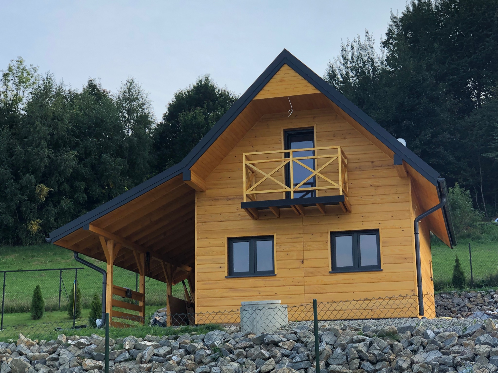 Żółty dom z drewna z czarnym dachem i małym balkonem, widok z dołu na tle drzew i skalniaka.