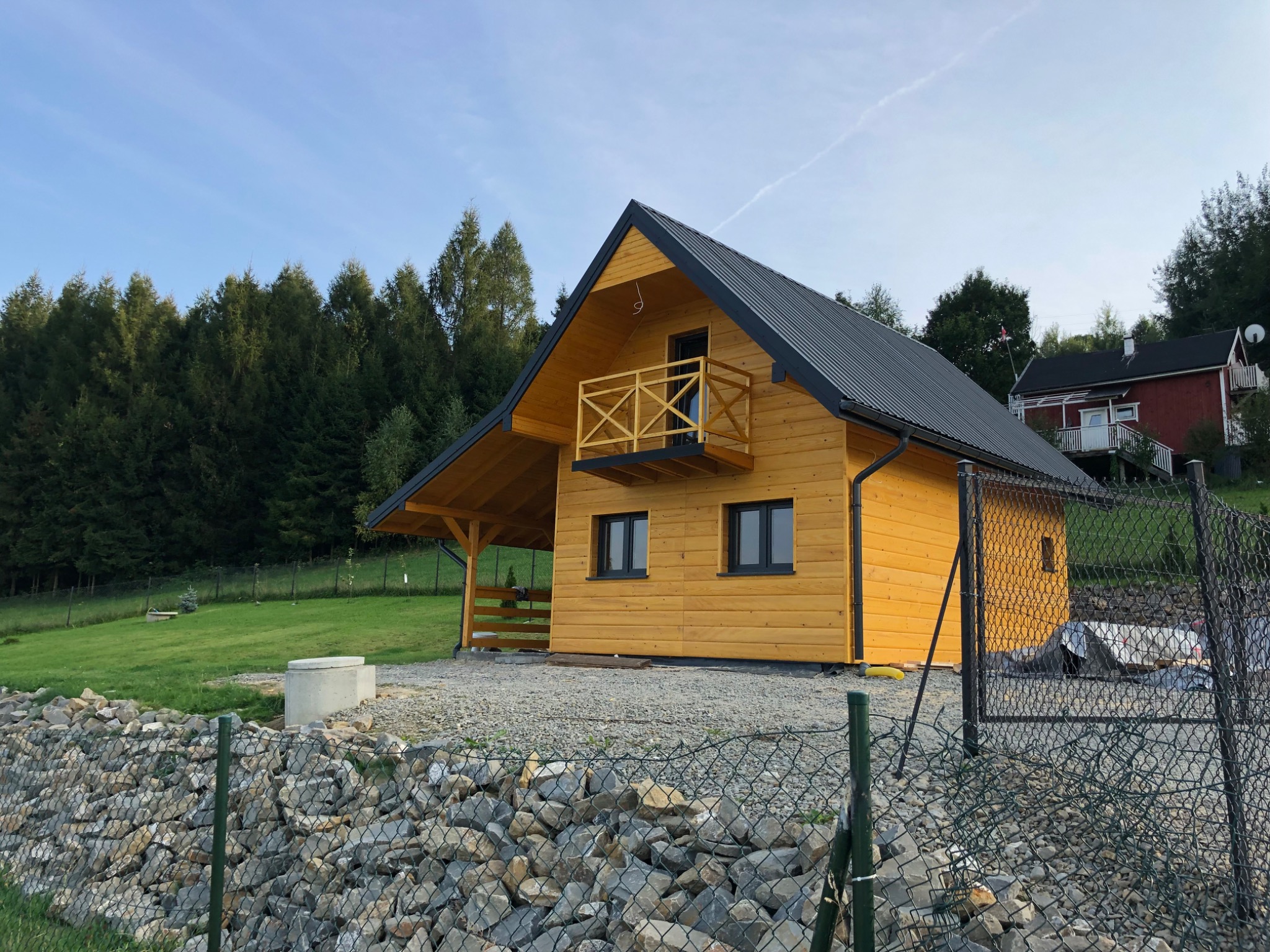 Nowoczesny, żółty dom z drewna z czarnym dachem i drewnianym balkonem, widoczny zza kamiennego murku i metalowej siatki ogrodzeniowej na tle lasu i błękitnego nieba.