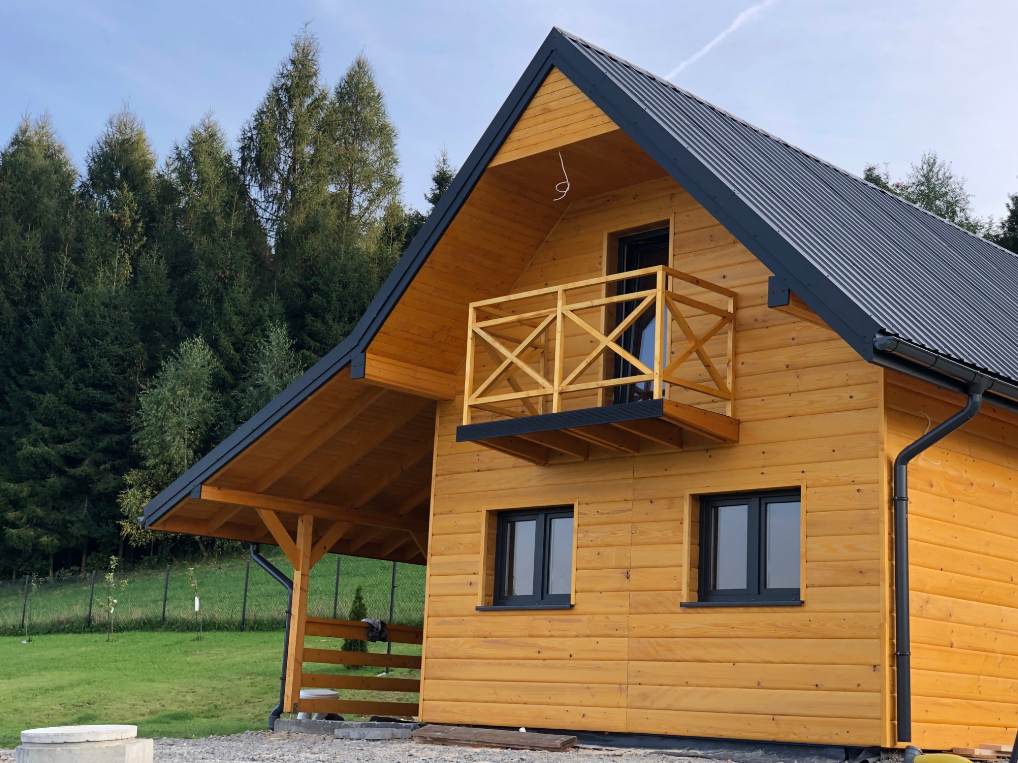 Jasny, drewniany dom z balkonem i ciemnym dachem na tle zielonego lasu i błękitnego nieba.