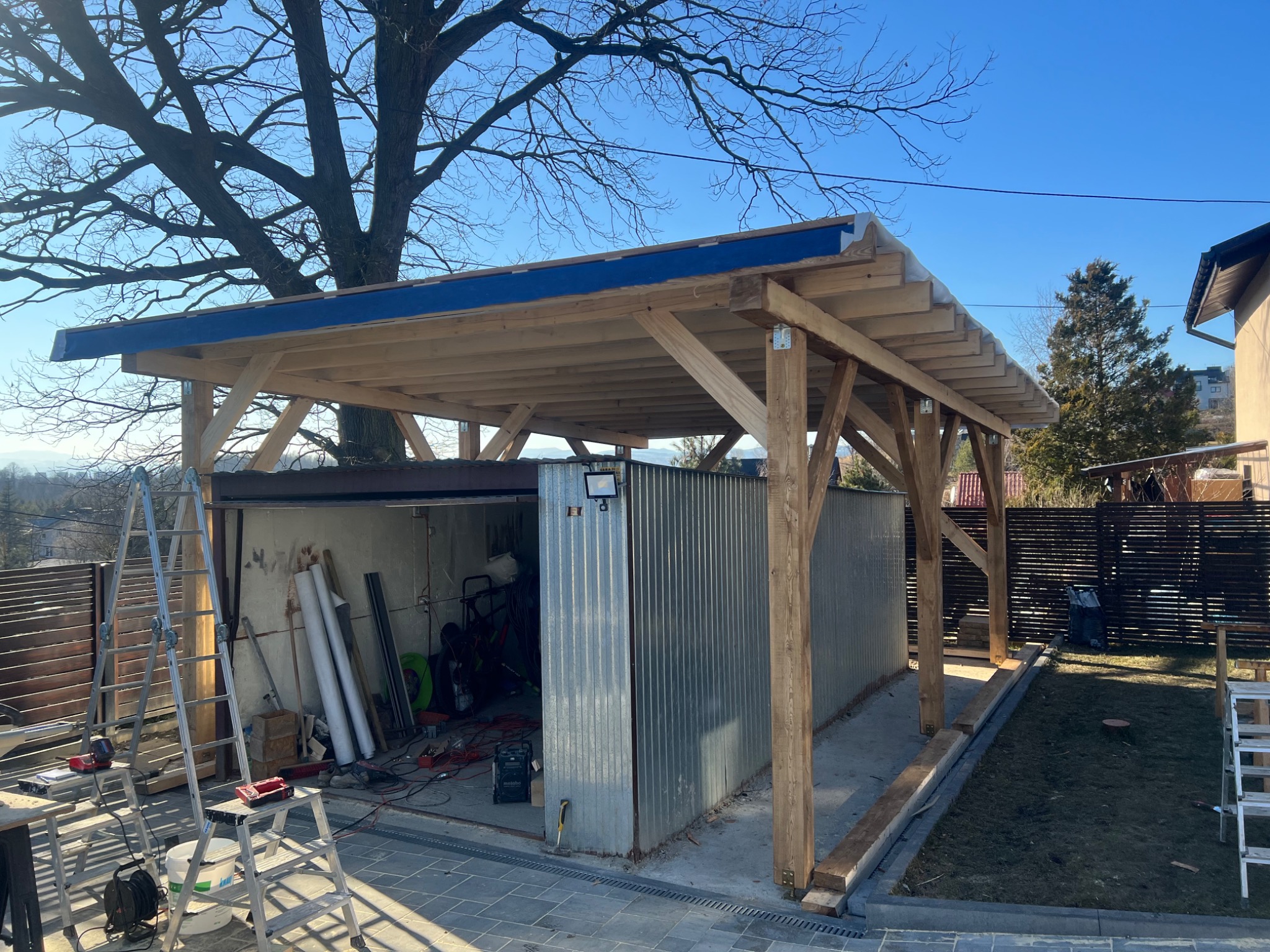 Konstrukcja z drewna, prawdopodobnie wiata garażowa w trakcie budowy, z widoczną metalową ścianą garażu i narzędziami w tle, na zewnątrz kostka brukowa i trawnik.