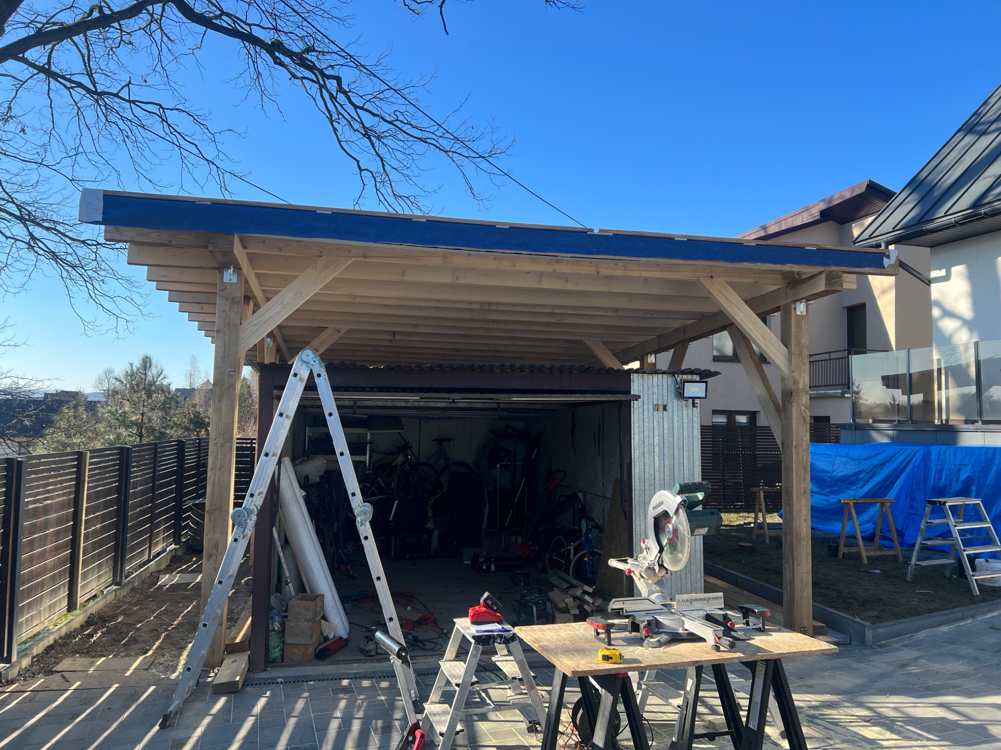 Konstrukcja drewnianego zadaszenia nad garażem w trakcie budowy, widoczna drabina, stół z narzędziami i piła do drewna na tle błękitnego nieba.
