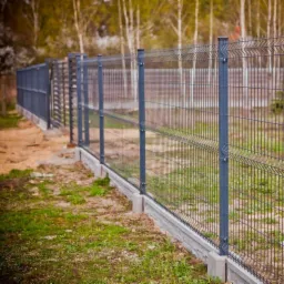 Szary panelowy system ogrodzeniowy na betonowej podmurówce, widok z boku, w tle drzewa.