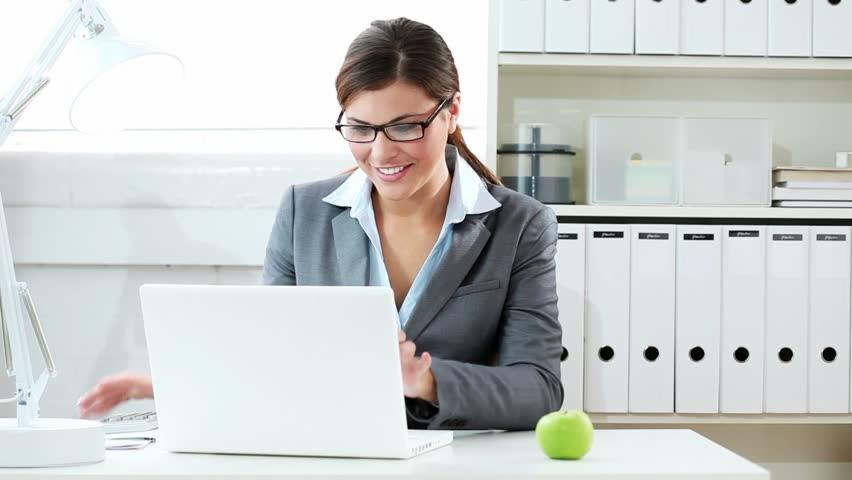 Kobieta w okularach i szarym żakiecie uśmiecha się pracując na laptopie przy biurku z jabłkiem, w tle regał z segregatorami.