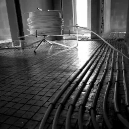 Czarno-białe zdjęcie przedstawia montaż ogrzewania podłogowego: rury ułożone na folii z rastrem, rozwijane z rolki na stojaku.