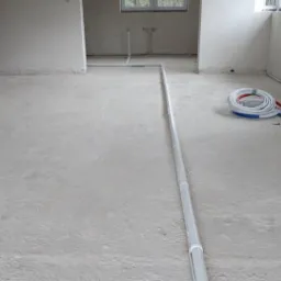 Wnętrze w stanie surowym z ułożonymi rurami instalacji grzewczej/hydraulicznej na podłodze, widoczne również zwinięte rury przy ścianie i okno w tle.