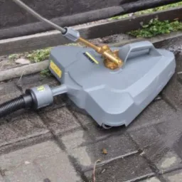 Szara myjka ciśnieniowa Karcher do powierzchni płaskich z widoczną dyszą i wężem, czyszcząca mokry, brudny brukowany taras z zielonymi resztkami w tle.