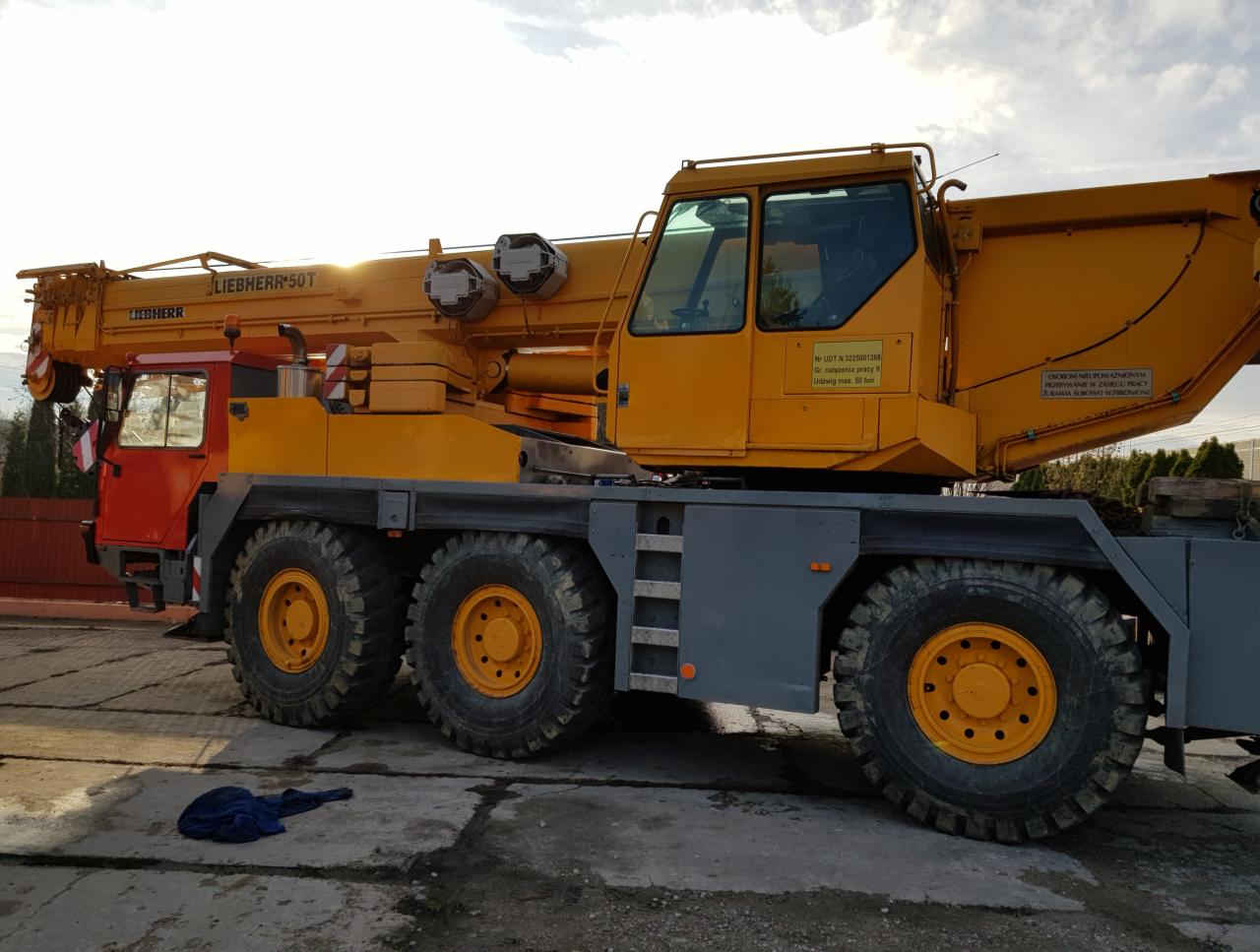 Żółto-czerwony dźwig samojezdny Liebherr 50T na ośmiu kołach, zaparkowany na betonowym placu, z widoczną tabliczką informacyjną na kabinie.