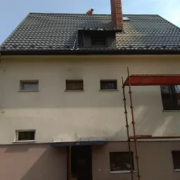 Dwupiętrowy budynek z ciemnoszarą, falistą blachodachówką, kominem z czerwonej cegły i drabina oparta o ścianę; na dachu widoczna postać robotnika.