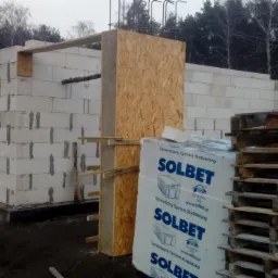 Wznoszenie ścian budynku z bloczków Solbet, widoczne szalunki i palety, teren budowy otoczony lasem.