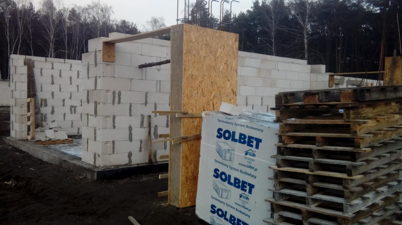 Wznoszenie ścian budynku z bloczków Solbet, widoczne szalunki i palety, teren budowy otoczony lasem.