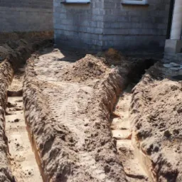 Wykopane rowy w piasku wokół budynku z szarej cegły, widoczne ślady opon na dnie rowu.