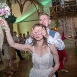 Panna młoda w koronkowej sukni z zasłoniętymi oczami przez mężczyznę, trzyma bukiet ślubny na sali balowej podczas wesela.