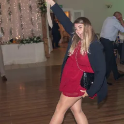 Rozradowana panna młoda w czerwonej sukience, trzymająca bukiet ślubny nad głową podczas weselnego przyjęcia w sali z drewnianą podłogą i dekoracjami świetlnymi.