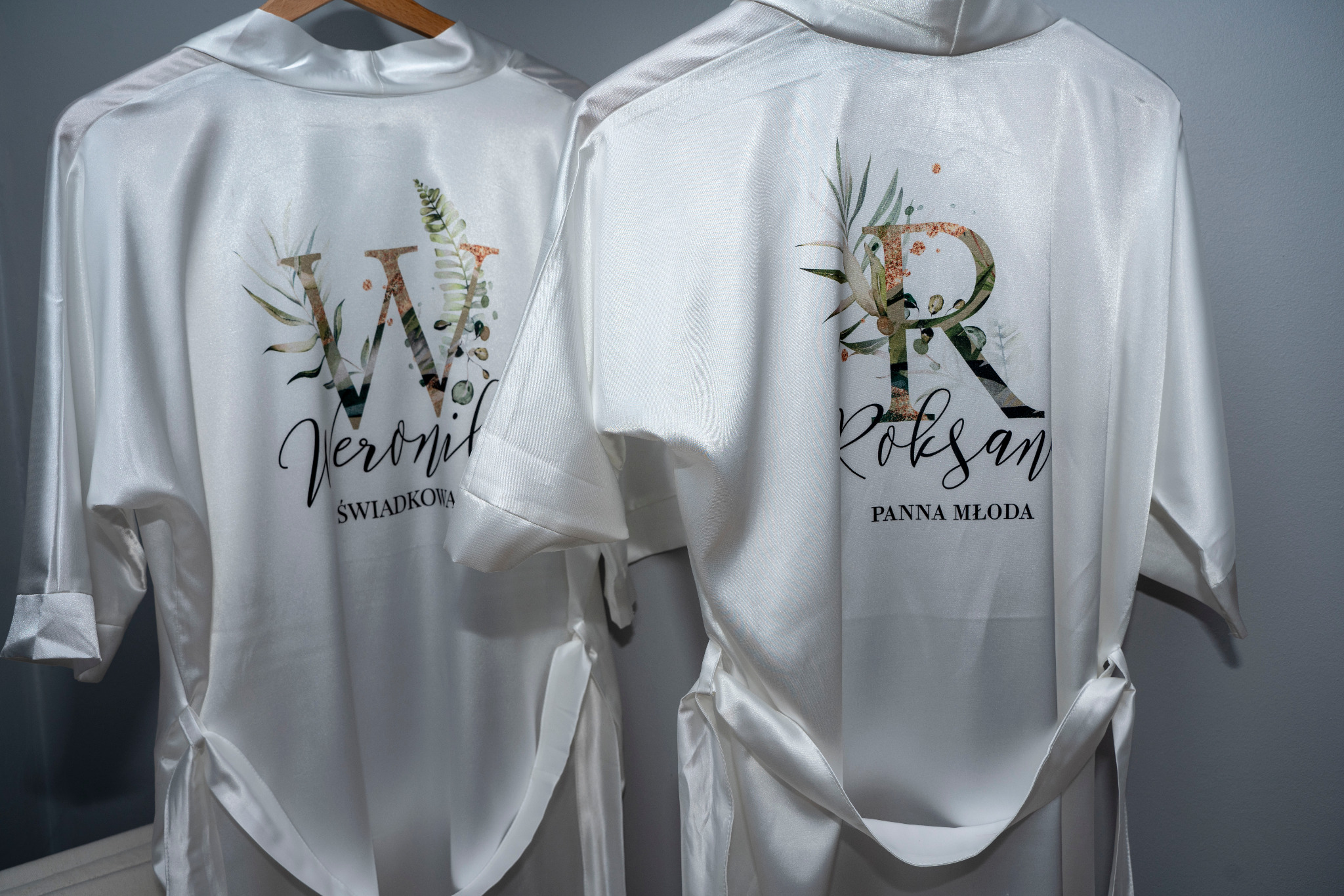 Dwa białe, satynowe szlafroki z nadrukowanymi imionami 'Weronika' i 'Roksana' oraz napisami 'Świadkowa' i 'Panna Młoda', ozdobione motywami roślinnymi, wiszące na wieszakach na tle szarej ściany.
