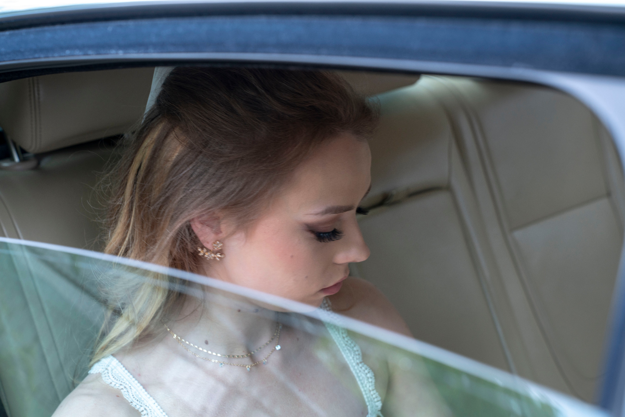 Panna młoda w sukni ślubnej, widoczna przez szybę samochodu, z delikatną biżuterią i eleganckim upięciem włosów.