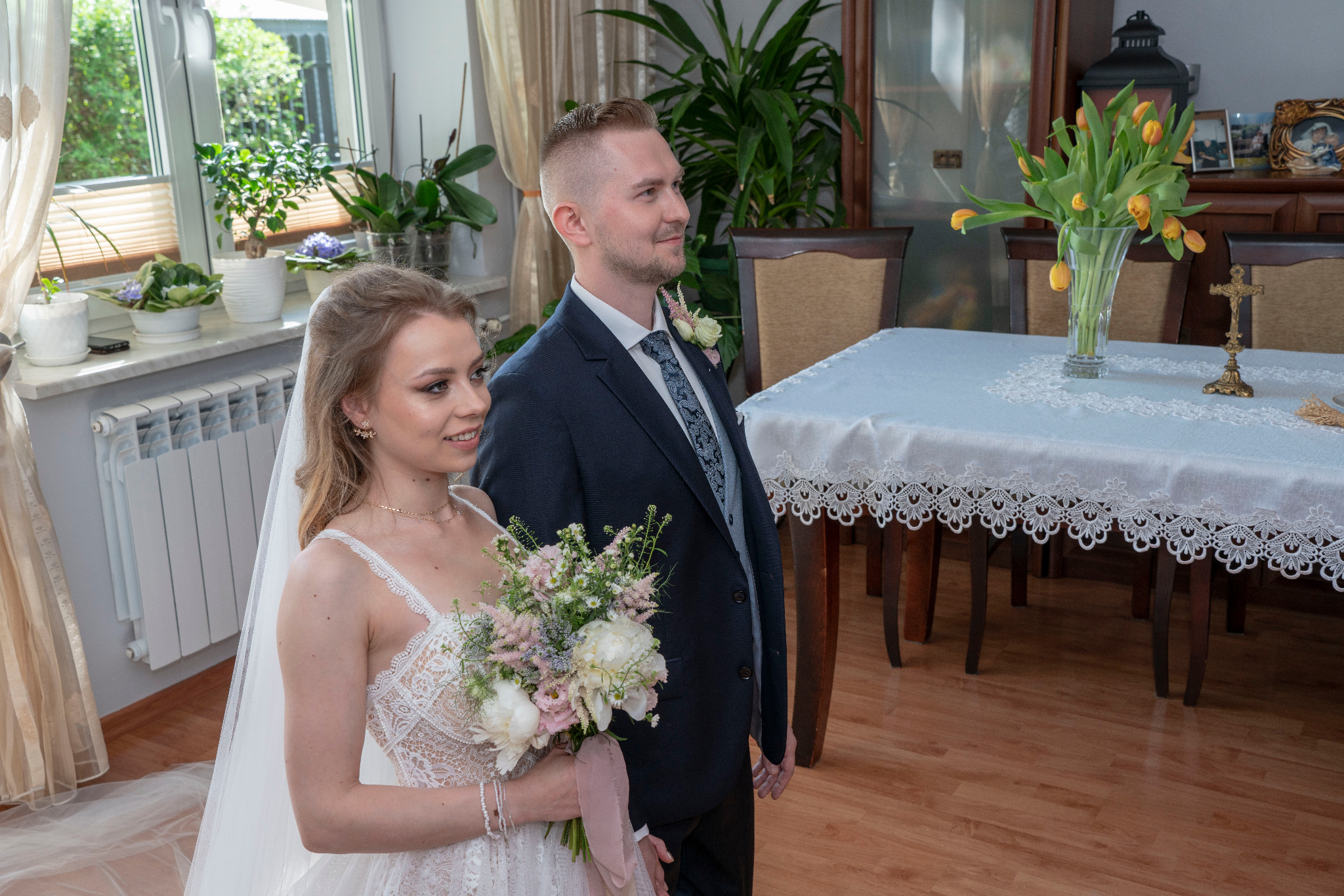 Młoda para w strojach ślubnych, panna młoda trzyma bukiet, w tle stół z tulipanami i dekoracjami.