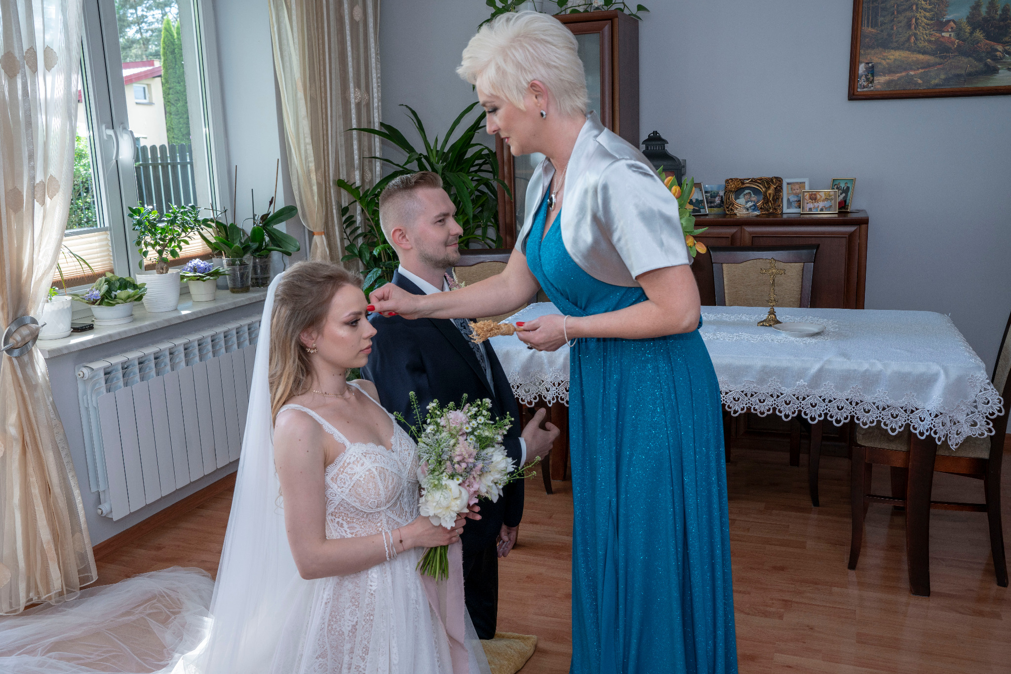 Błogosławieństwo młodej pary chlebem i solą podczas ceremonii powitania po ślubie, panna młoda w koronkowej sukni, pan młody w garniturze, kobieta w niebieskiej sukni z srebrnym szalem.