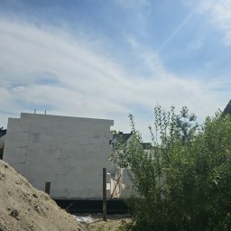 DomyJutra.pl Piotr Tereszkiewicz - Wznoszone ściany z białych bloczków betonu komórkowego na budowie domu, widoczna sterta piasku i zieleń krzewów na pierwszym planie, błękitne niebo w tle.